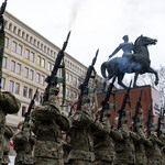 Żołnierze w mundurach polowych stoją w szeregu i wykonują salwę honorową, unosząc karabiny w górę. W tle znajduje się pomnik konny na wysokim cokole, a za nim fasada dużego budynku z licznymi oknami. Na niebie widoczny dym po wystrzałach.