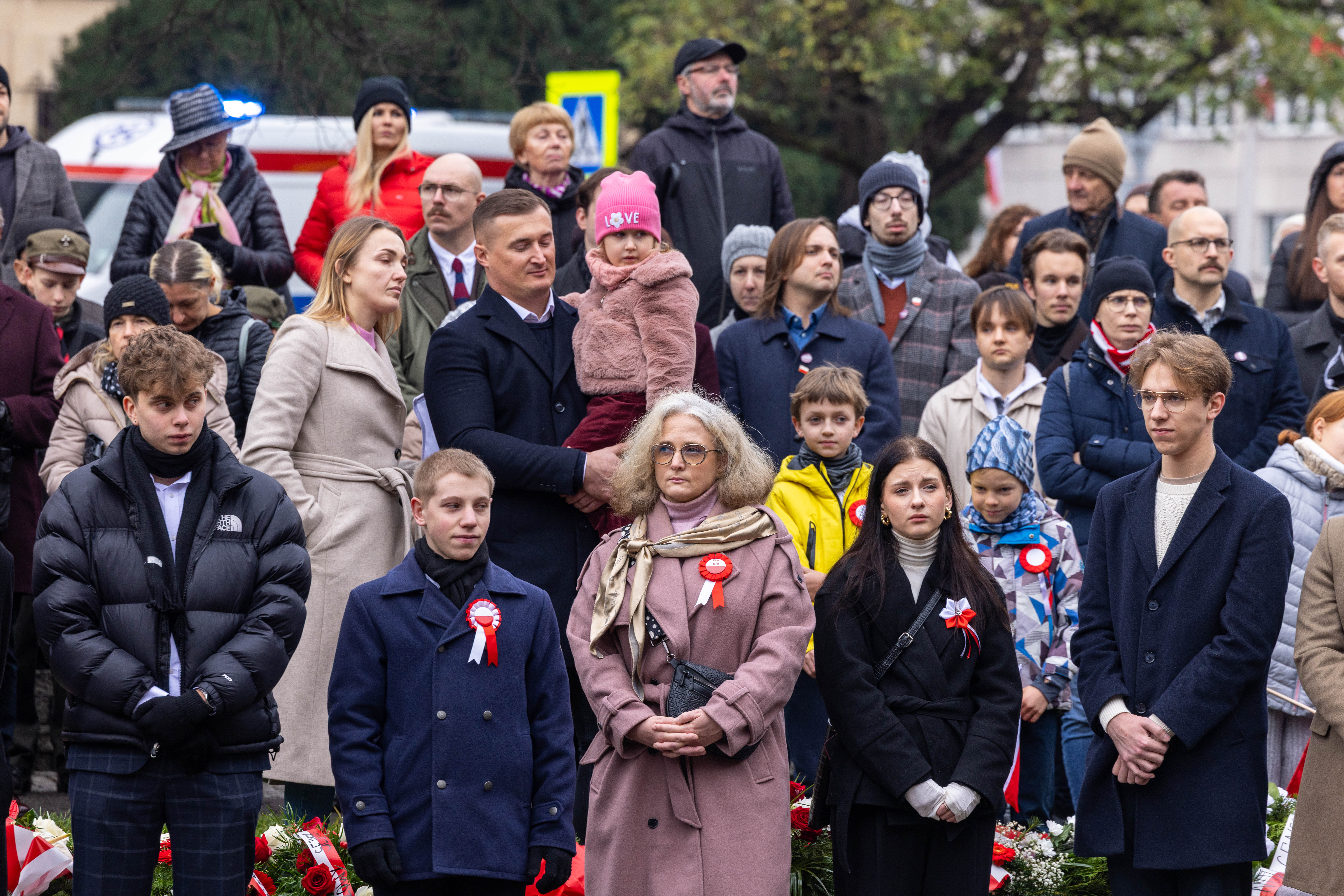 Grupa osób stoi w zwartym szyku podczas uroczystości patriotycznej. Uczestnicy ubrani są w eleganckie płaszcze i kurtki, część ma przypięte biało-czerwone kokardy. Na pierwszym planie widoczne są kwiaty ułożone w wieńce. W tle znajduje się pojazd służb oraz drzewa