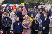 Grupa osób stoi w zwartym szyku podczas uroczystości patriotycznej. Uczestnicy ubrani są w eleganckie płaszcze i kurtki, część ma przypięte biało-czerwone kokardy. Na pierwszym planie widoczne są kwiaty ułożone w wieńce. W tle znajduje się pojazd służb oraz drzewa