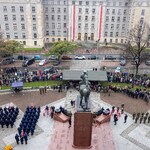 Widok z góry na plac przed monumentalnym budynkiem udekorowanym biało-czerwonymi flagami. Centralnie znajduje się pomnik konny na cokole, wokół którego zgromadzone są oddziały wojska w szyku, poczty sztandarowe oraz liczni uczestnicy uroczystości. Na ulicy stoją samochody, a wzdłuż chodników widoczne są grupy obserwatorów.
