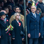 Grupa osób uczestniczy w oficjalnej uroczystości patriotycznej na świeżym powietrzu. Uczestnicy stoją w rzędzie, ubrani w eleganckie płaszcze, część z nich ma przypięte biało-czerwone kokardy. Jedna osoba trzyma bukiet kwiatów przewiązany wstążką w barwach narodowych. W tle widoczny tłum oraz elementy zadaszenia.