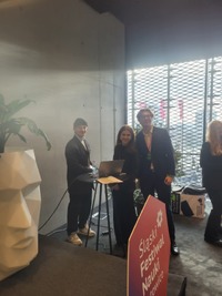 Stoisko Śląskiego Festiwalu Nauki w Katowicach; kilka osób stoi przy wysokim stoliku z laptopem, w tle duże okno z kratownicą, obok dekoracyjna donica w kształcie głowy z rośliną.