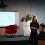 Prezentacja na temat zdrowia psychicznego i terapii odbywa się w nowoczesnej sali konferencyjnej. Na ekranie widoczny jest slajd z nagłówkiem „Zdrowie psychiczne” i dużym tytułem „Terapia”, obok którego znajdują się ikony dymków rozmowy. W pomieszczeniu stoją dwa czerwone fotele oraz mały stolik, a w tle znajduje się roślina doniczkowa. Kilka osób siedzi i słucha wystąpienia, a na stole z tyłu widoczny jest laptop.