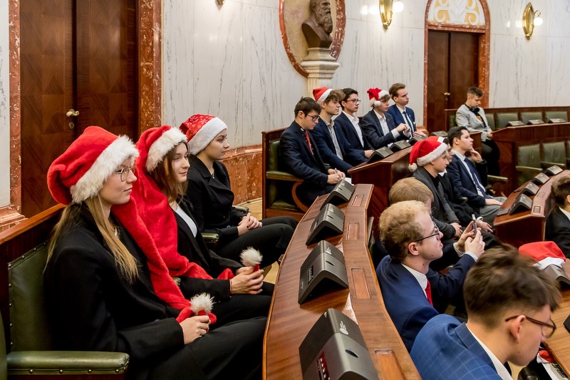 formalne spotkanie, prawdopodobnie w sali posiedzeń parlamentu lub rady miasta, charakteryzujące się eleganckimi drewnianymi biurkami, zielonymi skórzanymi krzesłami i marmurowymi ścianami z elementami dekoracyjnymi. W tle widoczna jest rzeźba przedstawiająca popiersie, która dodaje oficjalnego charakteru.
Kilka osób siedzi w rzędach, ubranych w formalne stroje, takie jak garnitury i marynarki, co wskazuje na wydarzenie zawodowe lub uroczyste. Uroczysty akcent jest widoczny, ponieważ wielu uczestników ma na sobie czerwone czapki Mikołaja, co sugeruje, że sesja odbywa się w okresie świątecznym.
Biurka mają wbudowane mikrofony i panele elektroniczne, typowe dla pomieszczeń parlamentarnych lub rad, co potwierdza, że jest to spotkanie o charakterze strukturalnym – zgodne z sesją parlamentu młodzieżowego lub sejmiku.
