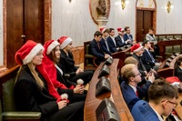 formalne spotkanie, prawdopodobnie w sali posiedzeń parlamentu lub rady miasta, charakteryzujące się eleganckimi drewnianymi biurkami, zielonymi skórzanymi krzesłami i marmurowymi ścianami z elementami dekoracyjnymi. W tle widoczna jest rzeźba przedstawiająca popiersie, która dodaje oficjalnego charakteru.
Kilka osób siedzi w rzędach, ubranych w formalne stroje, takie jak garnitury i marynarki, co wskazuje na wydarzenie zawodowe lub uroczyste. Uroczysty akcent jest widoczny, ponieważ wielu uczestników ma na sobie czerwone czapki Mikołaja, co sugeruje, że sesja odbywa się w okresie świątecznym.
Biurka mają wbudowane mikrofony i panele elektroniczne, typowe dla pomieszczeń parlamentarnych lub rad, co potwierdza, że jest to spotkanie o charakterze strukturalnym – zgodne z sesją parlamentu młodzieżowego lub sejmiku.