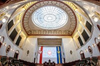 Wnętrze sali obrad parlamentu z półkolistym układem zielonych foteli, wysokimi marmurowymi ścianami oraz dużą kopułą ze szklanym sufitem ozdobionym ornamentami. Nad centralnym podium widoczne są flagi oraz godło państwowe, a całość oświetlona jest ciepłym światłem lamp rozmieszczonych wokół kopuły.