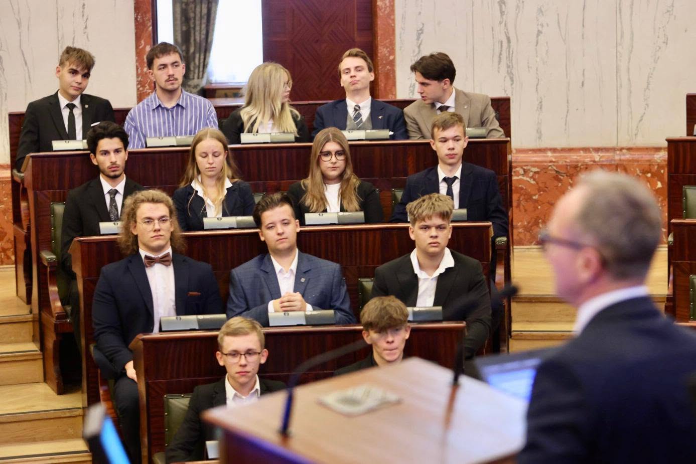 Uczestnicy sesji obrad siedzą w sali o półkolistym układzie ławek. Na pierwszym planie osoba trzyma dokument, a w tle widoczni są pozostali uczestnicy oraz marmurowe ściany z dekoracyjnymi elementami architektonicznymi