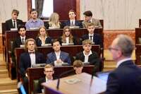 Uczestnicy sesji obrad siedzą w sali o półkolistym układzie ławek. Na pierwszym planie osoba trzyma dokument, a w tle widoczni są pozostali uczestnicy oraz marmurowe ściany z dekoracyjnymi elementami architektonicznymi