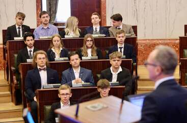 Uczestnicy sesji obrad siedzą w sali o półkolistym układzie ławek. Na pierwszym planie osoba trzyma dokument, a w tle widoczni są pozostali uczestnicy oraz marmurowe ściany z dekoracyjnymi elementami architektonicznymi