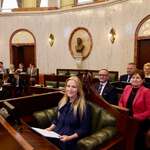 Grupa młodych uczestników zajmuje miejsca na widowni sali obrad. Na pierwszym planie widoczna jest mównica oraz osoba przemawiająca, uchwycona nieostro od tyłu